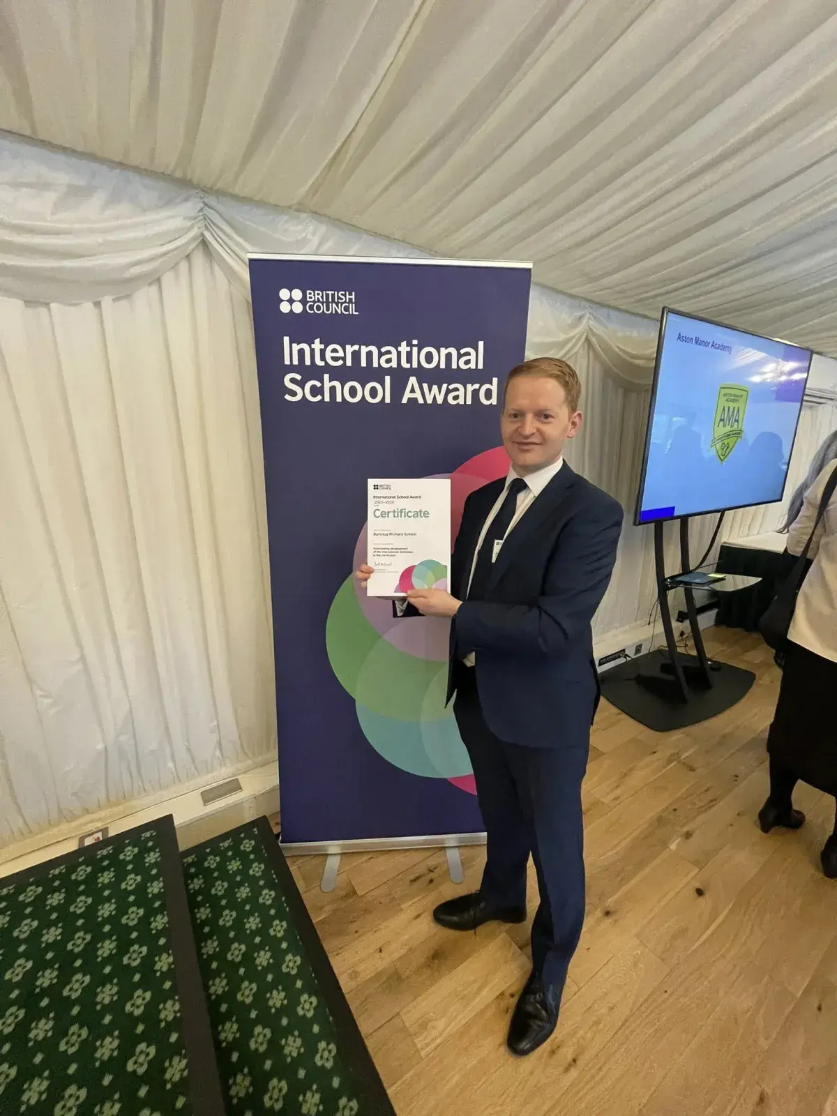 Dunclug PS Principal holding Award standing in front of a British Council pop up